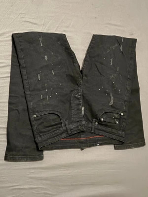 Svarta slim jeans  - Säljer ett par svarta slim fit jeans med slitningar och distressed detaljer. Jeansen har fem fickor, knappgylf och röda sömmar på insidan. Tillverkade i Italien och har en modern, trendig look.