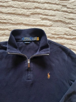 Marinblå halvzip från Polo Ralph Lauren - Klassisk marinblå tröja från Polo Ralph Lauren med halvzip och hög krage. Tröjan har lång ärm, ribbade muddar och den ikoniska broderade loggan på bröstet. Perfekt till jeans eller chinos för en clean och stilren look. Inga skavanker. Längd 63cm. Bredd: 52cm