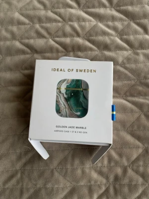 AirPods-fodral Golden Jade Marble - Snyggt AirPods-fodral från iDeal of Sweden i mönstret Golden Jade Marble. Fodralet har en grön, vit och guldmarmorerad design och passar till AirPods 1:a och 2:a generationen. Tillverkat i hårdplast med gulddetaljer och stilren finish.