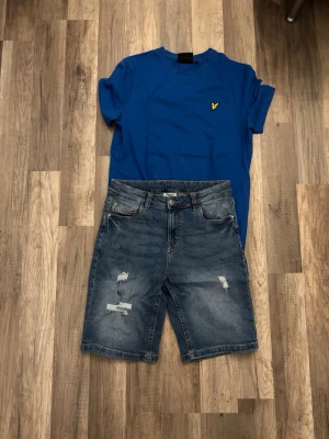 Blå jeansshorts med slitningar - Säljer ett par blå jeansshorts med slitna detaljer och klassisk femficksdesign. Med en mörkblå lyle Scott t-shirt som matchar dom blåa shortsen
