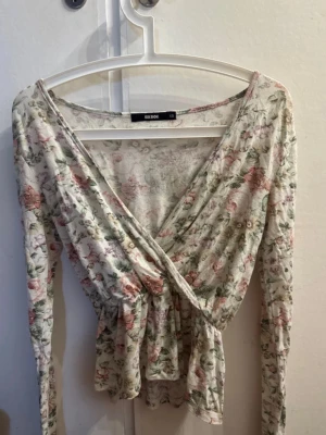 Blommig omlottopp från Bik Bok - Supersöt omlottopp från Bik Bok i blommigt mönster med pastelliga toner av rosa, grön och beige. Toppen har v-ringning, lång ärm och en lätt volang nertill. Perfekt för dig som gillar romantisk och feminin stil.