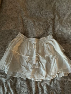 Vit kortkjol med volang och snörning - Supersöt vit kortkjol med volang och dekorativ snörning med tofsar och pärlor i midjan. Kjolen har en luftig och lätt känsla, och den är stretchig 🤍.