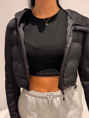 Svart croppad pufferjacka med huva - Säljer en svart croppad pufferjacka med huva och dragkedja framtill. Jackan har quiltad design på kroppen och släta ärmar, vilket ger en sportig och trendig look. Perfekt för att hålla värmen under kyliga dagar och passar till många olika outfits.