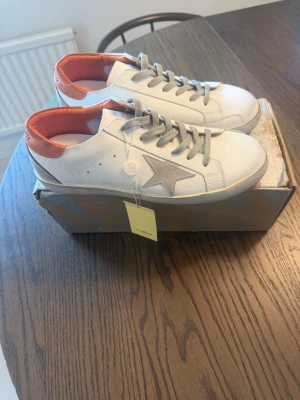 Golden Goose Superstar - Snygga vita sneakers med grå stjärna på sidan och grå skosnören. Insidan är fodrad med orange detalj runt hälen. Skorna har en klassisk låg profil och är tillverkade i slätt läderliknande material. Perfekta för en trendig och avslappnad stil.