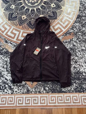 Svart träningsjacka från Nike Miller  - Snygg svart träningsjacka från Nike med dragkedja framtill och huva. Jackan har vita detaljer och Nike-logga på bröstet. Tillverkad i lätt och funktionellt material som passar perfekt för löpning eller annan träning.