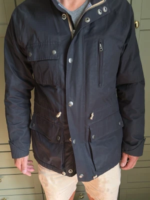 Mörkblå parkas med huva och fickor - Snygg mörkblå parkas med flera stora fickor och dragkedja samt tryckknappar framtill. Jackan har justerbar midja med snören och en praktisk huva. Materialet ser ut att vara slitstarkt och vattenavvisande, perfekt för kyligare dagar.