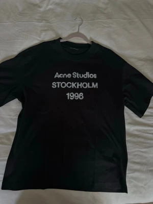 Svart Acne Studios t-shirt - Säljer en svart t-shirt Acne Studios med tryck i vitt på bröstet där det står 'Acne Studios STOCKHOLM 1996'. Klassisk passform och rund halsringning. Perfekt för dig som gillar stilrena och ikoniska plagg. Obs inte äkta