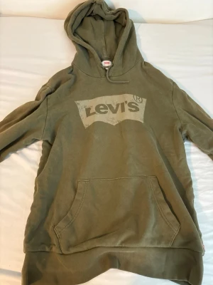 Olivgrön hoodie från Levi's - Säljer en olivgrön hoodie från Levi's med stor logga framtill. Hoodien har huva med dragsko och en stor magficka. Tillverkad i mjukt bomullsmaterial, perfekt för chill dagar. Passar dig som gillar en avslappnad och stilren look.