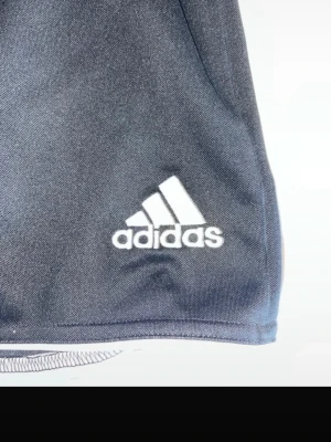 Mörkgrå Adidas shorts i polyester - Snygga mörkgrå shorts från Adidas med klassisk vit logga på benet. Tillverkade i lätt och funktionell polyester med CLIMALITE-teknologi som andas. Perfekta för träning eller chill dagar. Enkel design utan extra detaljer.