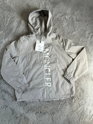 Beige grå vindjacka från Moncler - Snygg beige grå vindjacka från Moncler med huva och dragkedja framtill. Jackan har stora vita Moncler-bokstäver längs dragkedjan, två fickor med dragkedja och justerbar nederkant. Tillverkad i lätt, vindtätt material och har Moncler-logga på ärmen. Enda problemet med jackan är att logan på armen är lite trasig därför säljs den för så billigt 