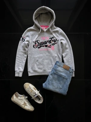 Grå hoodie från Superdry med tryck - Säljer en grå hoodie från Superdry med svart och rosa tryck på bröstet och siffran 26 på ärmen. Hoodien har en stor huva med dragsko, magficka och mjukt fleecefoder. Perfekt för en avslappnad och sportig stil. Plagget är använt men i riktigt fint skick. Perfekt för dig som vill sticka ut inför skolstarten. Markerad med storlek L men passar för S/M. Priset är ej hugget i sten.