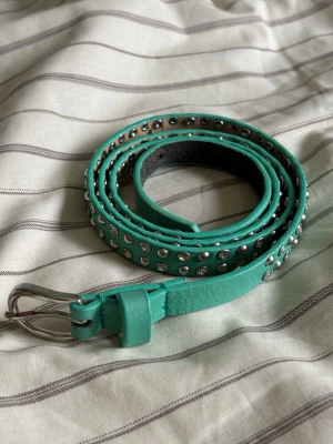 Turkost bälte med nitar - Säljer ett smalt turkost bälte med silverfärgade nitar längs hela bandet. Bältet har ett klassiskt spänne i metall och är gjort i ett material som liknar skinn. Perfekt för att ge din outfit en cool touch.