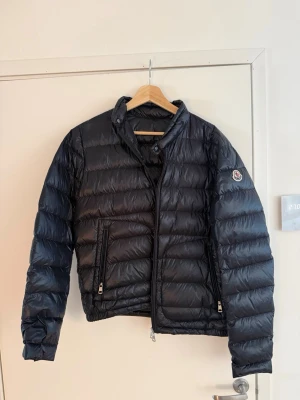 Moncler jacka | storlek 3 - Säljer en marinblå Moncler dunjacka i storlek 3. Sparsamt använd och i mycket fint skick.  Jackan är hel och fräsch utan hål, revor eller andra skador. Dragkedjor och fickor fungerar som de ska. Klassisk Moncler-design med blankt marinblått yttertyg, dunfyllning och logotyp på ärmen.  • Märke: Moncler • Storlek: 3 • Färg: mörk marinblå • Skick: mycket fint begagnat skick • Sparsamt använd • Inga hål eller skador  Fler bilder på etiketter, detaljer och mått kan skickas vid seriöst intresse.