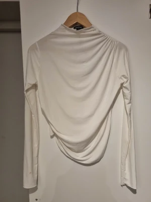 Vit långärmad topp från Boohoo (fläck finns) - Säljer en vit långärmad topp från Boohoo med draperad rygg och hög hals. Toppen har en elegant och stilren look, perfekt för dig som gillar trendiga detaljer. Tillverkad i ett mjukt och stretchigt material som sitter snyggt på kroppen.