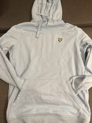Ljusblå hoodie från Lyle & Scott - Snygg ljusblå hoodie från Lyle & Scott med klassisk gul logga på bröstet. Hoodien har känguruficka, dragsko i huvan och ribbade muddar. Perfekt för en avslappnad och stilren look.