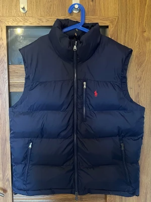 Polo Ralph Lauren väst - Snygg mörkblå dunväst från Polo Ralph Lauren med hög krage, dragkedja framtill och tre fickor med dragkedja. Klassisk röd logga på bröstet och quiltad design. Perfekt för lager på lager och streetwear-vibe. Använt max en gång, priset kan diskuteras kom pm.