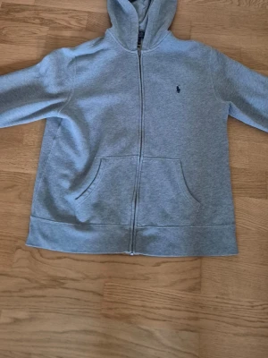 Ljusgrå hoodie med dragkedja - Ljusgrå hoodie med dragkedja och huva. Klassisk design med känguruficka framtill och en liten mörkblå logga broderad på bröstet. Tillverkad i mjukt sweatshirtmaterial som är skönt att ha på sig. Perfekt för en avslappnad stil.
