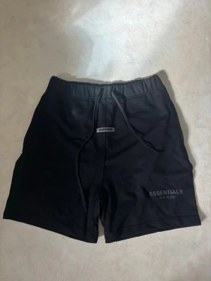 Svarta shorts Essentials Fear of God - Säljer ett par svarta shorts från Essentials Fear of God med snörning i midjan och logga tryckt på vänster ben. De har en avslappnad passform och är perfekta för sommaren. Materialet känns mjukt och bekvämt, troligen bomull eller bomullsblandning. Storlek L ej använda