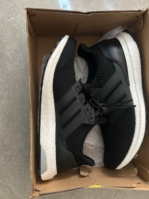 Svarta Adidas Ultraboost sneakers - Snygga svarta Adidas Ultraboost sneakers med stickad ovandel och klassiska tre ränder på sidan. Skorna har vit Boost-mellansula och svart yttersula från Continental. Perfekta för dig som gillar stilrena och bekväma sneakers.