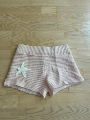 Ljusrosa stickade shorts med stjärndetalj - Säljer ett par ljusrosa stickade shorts med bred resår i midjan och en vit stjärna broderad på ena benet. Shortsen har en mjuk och bekväm känsla, perfekta för varma dagar eller mys hemma. Köpte på Plick men dom är från 