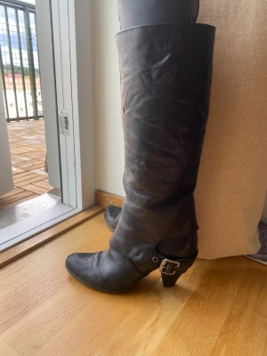 Svarta höga läderstövlar - Säljer ett par svarta höga stövlar i läder med rundad tå och dekorativt spänne vid ankeln. Klacken är låg och stabil. Dom har varit väldigt sköna men har blivit för små😭