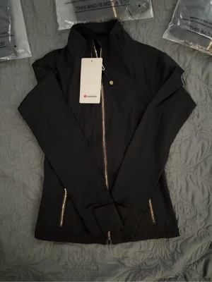 Svart träningströja från Lululemon - Snygg svart tränings jacka från Lululemon med hög krage och guldfärgade dragkedjor. Jackan har figurnära passform, långa armar och två fickor framtill. Perfekt för dig som vill ha en sportig och stilren look.