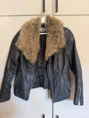 Svart skinnjacka med fuskpäls Jofama - Snygg svart skinnjacka från Jofama by Kenza i storlek 34. Jackan har en stor, brun fuskpäls-krage och dragkedjor på fickor och ärmslut. Perfekt för dig som vill ha en edgy look med lite extra attityd.