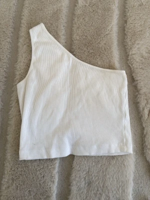 Vit oneshoulder topp från Even&Odd - Säljer en vit ribbad oneshoulder topp från Even&Odd. Toppen är ärmlös och har en asymmetrisk axel, vilket ger en trendig look. Materialet är mjukt och stretchigt, perfekt för att styla med jeans eller kjol.