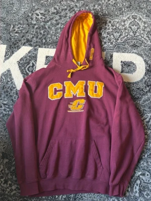 Vinröd Hoodie med gul CMU-text - Säljer en vinröd hoodie med stor gul CMU-text och logga på bröstet. Hoodien har gul insida i huvan och matchande gula snören. Klassisk känguruficka framtill och mjukt material, perfekt för chill dagar.