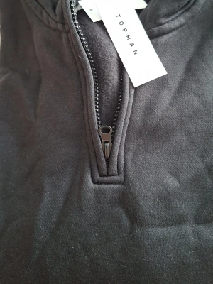 Svart hoodie med dragkedja från Topman - Snygg svart hoodie från Topman med halv dragkedja framtill och ribbade muddar vid ärmsluten. Tillverkad i mjukt bomullstyg och har en klassisk huva. Perfekt för en avslappnad och stilren look.