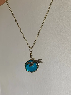 Halsband med blå sten och trollslända - Unikt halsband med en rund, klarblå sten infattad i en guldfärgad ram. Hängsmycket pryds av en detaljrik trollslända i metall. Kedjan är guldfärgad och har en klassisk länkdesign. Perfekt för dig som gillar boho eller vintageinspirerad stil.