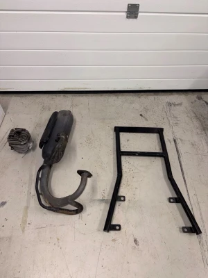 Slider 12:a - Säljer ett svart mopedram, en cylinder i metall samt ett avgassystem med ljuddämpare och böjt rör. Perfekt för dig som bygger eller renoverar moped. Alla delar är av metall och har klassisk design för äldre mopeder.