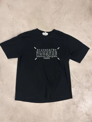 Maison Margiela svart t-shirt - Svart t-shirt från Maison Margiela med ikoniskt tryck framtill i vitt med siffror och logga. Klassisk passform och rund halsringning. Storleken syns inte men skulle säga large, är 188, 85 kg den sitter bra! Sparsamt använd