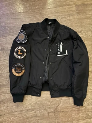 Svart bomberjacka med patches - Svart bomberjacka med quiltat foder och flera patches på ena ärmen. Jackan har detaljer med texten LCKR på bröstet.