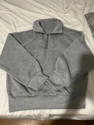 Grå halvzip hoodie i sweatshirtmaterial - Säljer en stilren grå hoodie med halv dragkedja och hög krage. Tillverkad i mjukt sweatshirtmaterial med ribbade muddar vid ärmar och nederkant. Perfekt för dig som gillar en clean och avslappnad look.