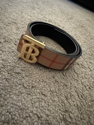 Stilig lysande burberry bälte  - Snyggt bälte i beige med klassiskt rutigt mönster och en stor guldfärgad spänne med bokstäver. Perfekt accessoar för att lyfta din outfit och ge en exklusiv touch. Bältet har svarta kanter och insida.