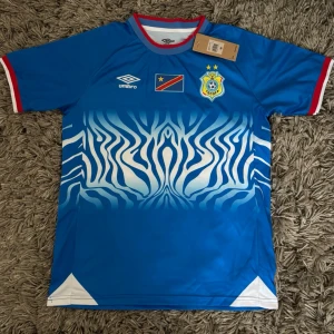 Blå Umbro fotbollströja DR Kongo - Säljer en blå fotbollströja från Umbro med DR Kongos flagga och förbundsmärke på bröstet. Tröjan har ett vitt och blått zebramönster framtill, röda detaljer vid krage och ärmslut samt vita trianglar vid nederkanten. Perfekt för match eller träning!