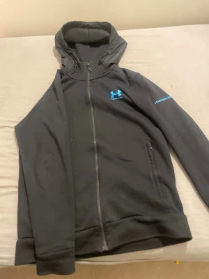 Svart hoodie från Under Armour - Säljer en svart hoodie från Under Armour med dragkedja och blå logga på bröstet samt tryck på ärmen. Hoodien har huva och två fickor framtill. Perfekt för dig som gillar sportig och clean stil.