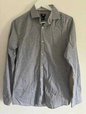 Grå skjorta från H&M - Klassisk grå skjorta från H&M i bomull med vit knappslå och diskreta detaljer på kragen. Skjortan har lång ärm och en stilren, enkel design som passar till många olika looks.