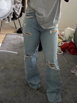 Ljusblå slitna raka jeans - Säljer ett par ljusblå jeans med raka ben och flera slitningar och hål på både fram- och baksida. Jeansen har hög midja och klassisk femficksmodell. Perfekta för en avslappnad och trendig look.