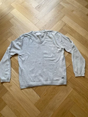 Ljusgrå stickad tröja  - Ljusgrå stickad tröja med V-ringning och långa ärmar. Tröjan har ribbade muddar vid ärmslut och nederkant samt en liten metallbricka vid nederkanten. Enkel och stilren design, perfekt för lager på lager.