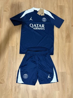 PSG träningsset i mörkblått - Snyggt träningsset från PSG med matchande t-shirt och shorts i mörkblått med vita detaljer. Setet har tryckta loggor från Paris Saint-Germain och Jordan samt Qatar Airways på tröjan. Tillverkat i ett lätt och ventilerande funktionsmaterial, perfekt för fotbollsträning.