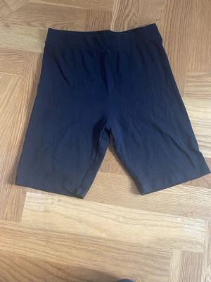 Svarta cykelshorts i bomull - Ett par svarta cykelshorts med midja i stretchig bomull. De har en enkel och clean design utan synliga detaljer eller mönster, perfekta för en avslappnad och sportig stil. Passformen är normal och materialet känns mjukt mot huden.