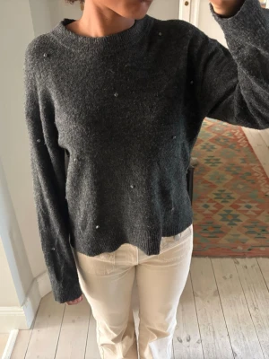 Mörkgrå stickad tröja med pärlor - Mysig mörkgrå stickad tröja med små pärldetaljer över hela tröjan. Tröjan har rund halsringning, långa ärmar och ribbade muddar vid ärmslut och nederkant. Perfekt för dig som vill ha en enkel men ändå unik look.