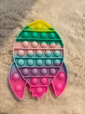 Regnbågsfärgad pop it raket - Säljer en pop it i form av en raket med regnbågsfärger i grönt, gult, rosa, blått och lila.