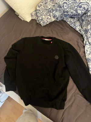 Svart sweatshirt från Moncler - Svart sweatshirt från Moncler med rund halsringning och diskret Moncler-logga på bröstet. Tröjan har ribbade muddar vid ärmslut och nederkant samt en liten rödvit detalj i nacken. Perfekt för en stilren och enkel look. Aldrig använd ute.