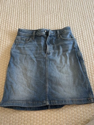 Blå jeanskjol från Lee - Klassisk blå jeanskjol från Lee med fem fickor, bälteshällor och knappgylf. Kjolen har en rak passform och slutar ovanför knät. Perfekt till vardags för en avslappnad och trendig look.