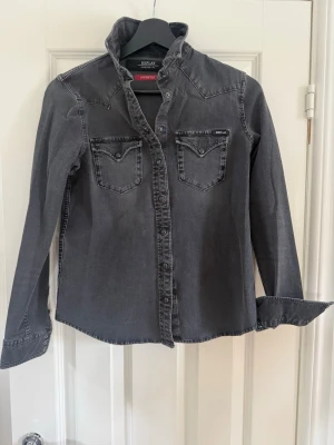 Grå jeansskjorta från Replay - Snygg grå jeansskjorta från Replay med tryckknappar och två bröstfickor. Skjortan har lång ärm och klassisk krage. Materialet är slitstark bomull med en tvättad look. Perfekt för en avslappnad och cool stil. Passar mig som är storlek 36/38 