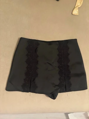 Svart kortkjol med spetsdetaljer - Snygg svart kortkjol i satinliknande material med två breda spetsdetaljer framtill. Kjolen har en rak passform och är perfekt för dig som gillar en stilren men ändå trendig look.