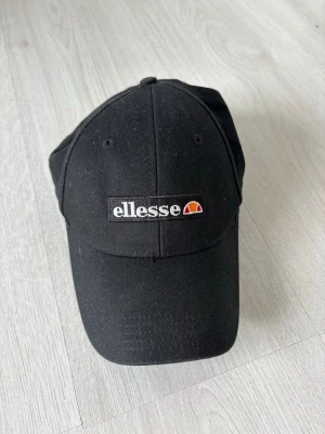 Svart keps från Ellesse - Snygg svart keps från Ellesse med broderad logga framtill. Klassisk böjd skärm och justerbar rem baktill. Tillverkad i bomullstyg och har ventilerande hål upptill för extra komfort. Perfekt accessoar för en sportig och trendig look.